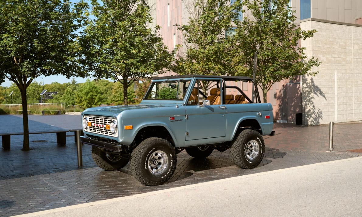 Brittany Blue 1966 Ford Bronco with Whiskey leather interior, parked curbside in an urban setting. Vintage Bronco restoration featuring Gen 3 Coyote 5.0L engine, lifted stance, and chrome accents.