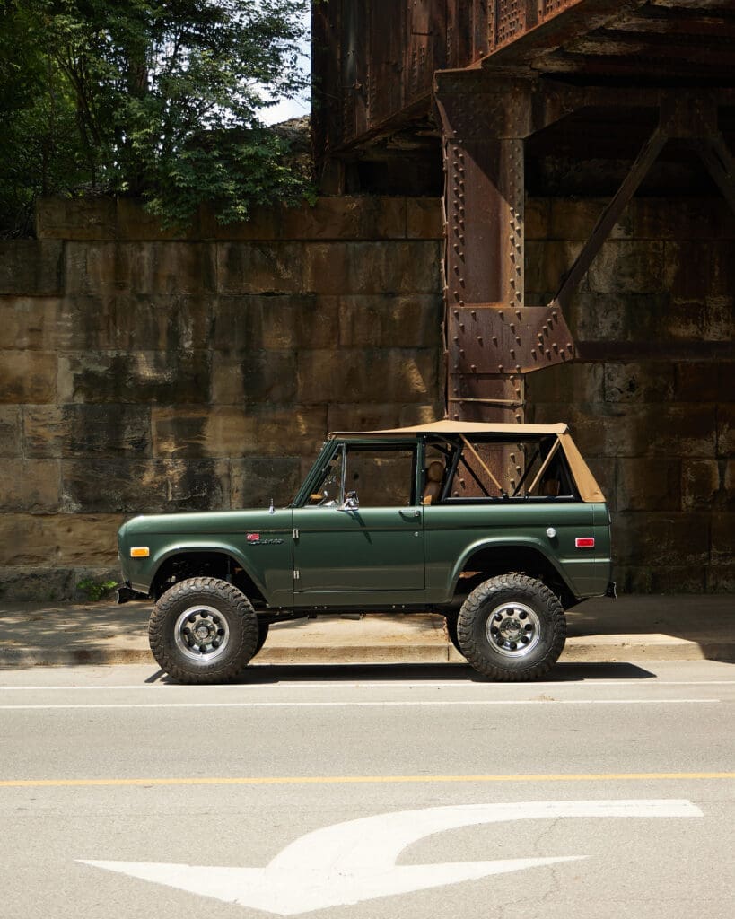 A 1967 Classic Ford Bronco named “Walloon Lake” is parked along a city street beneath a rusted steel bridge, highlighting its lifted stance, tan soft top, and meticulous restoration. Built by Classic Ford Broncos, this vintage SUV blends rugged performance with refined craftsmanship and timeless design.