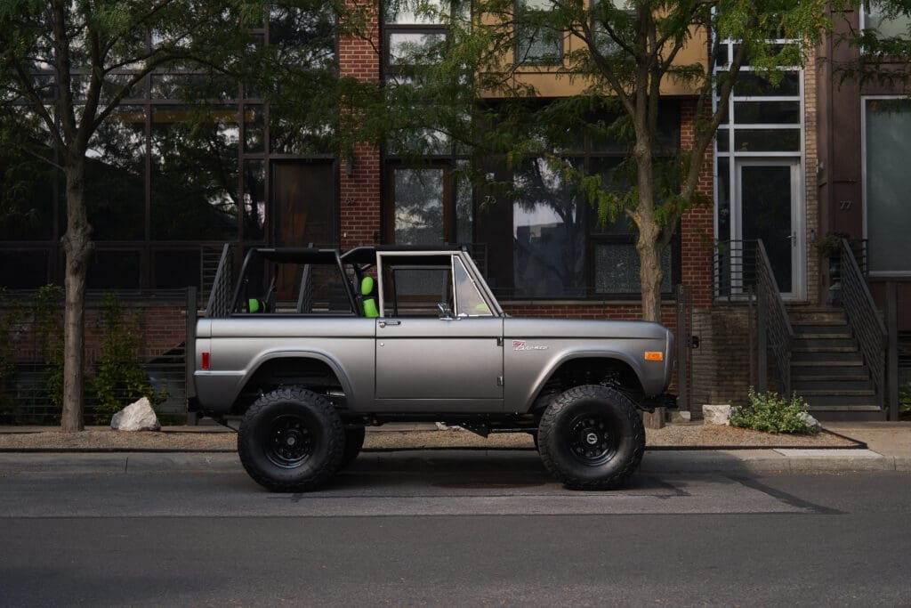 A 1977 Classic Ford Bronco named “Green Monster” is parked on a city street, showcasing its lifted stance, open cabin, and modernized design. Built by Classic Ford Broncos, this custom SUV combines vintage styling with a Gen 3 Coyote 5.0L engine, advanced suspension, and handcrafted interior for the ultimate blend of power and refinement.