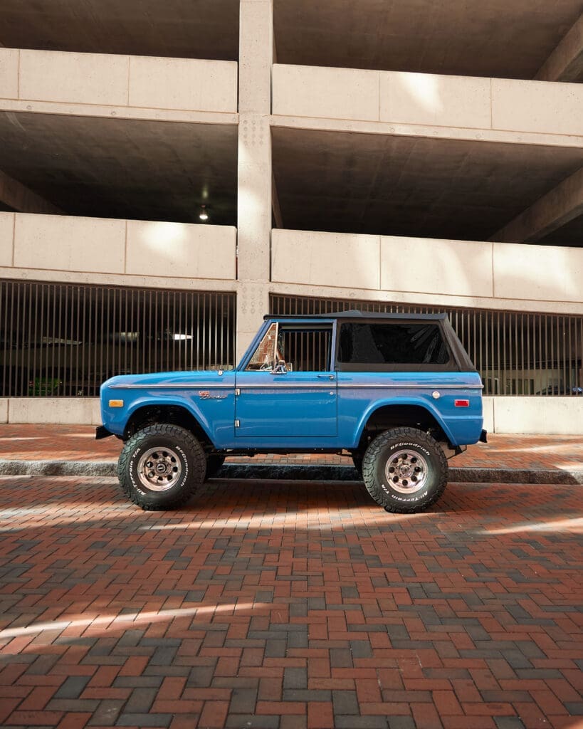 Restored 1974 Classic Ford Bronco in Bahama Blue with a black soft top, lifted suspension, and BFGoodrich all-terrain tires parked in front of a modern concrete parking structure. This vintage Ford Bronco combines timeless design with upgraded off-road performance, showcasing Classic Ford Broncos’ expert craftsmanship and attention to detail.