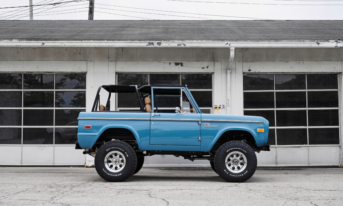 1973 Classic Ford Bronco parked in front of an old industrial garage with lifted suspension, black roll cage, and BFGoodrich All-Terrain tires. This restored vintage Ford Bronco highlights Classic Ford Broncos’ signature craftsmanship—combining modern performance, off-road capability, and authentic first-generation design.