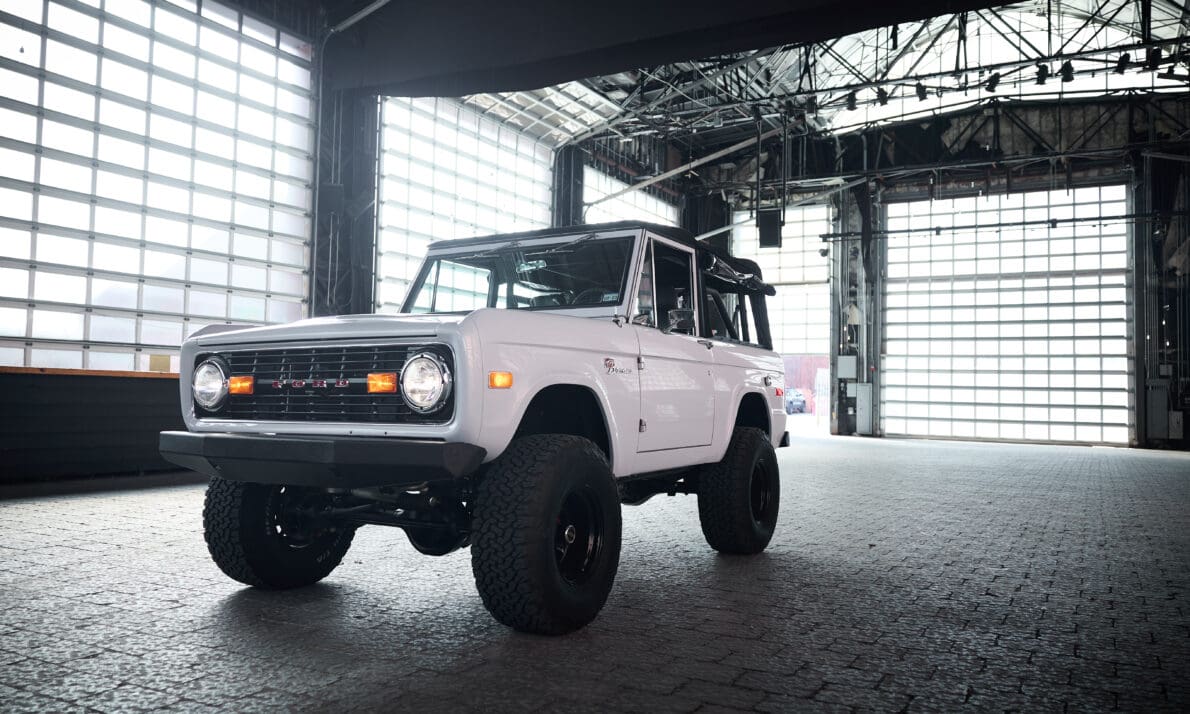 Restored 1975 Classic Ford Bronco in Yulong White with a black soft top, lifted suspension, and off-road tires parked inside an industrial warehouse with large glass doors. This vintage Ford Bronco exemplifies Classic Ford Broncos’ craftsmanship, blending modern performance upgrades with the timeless design of the first-generation Bronco.
