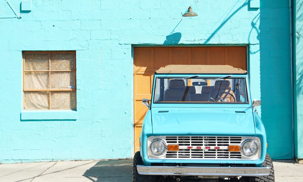 Front view of restored Classic Ford Bronco in Peacock Blue with tan soft top parked in front of a turquoise building, vintage early Bronco featuring lifted suspension, off-road tires, and custom restoration by Classic Ford Broncos.