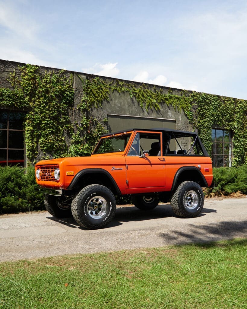 Classic Ford Bronco in bright orange with black soft top, lifted suspension, and off-road tires parked in front of an ivy-covered building, showcasing vintage 4x4 styling and restored details.