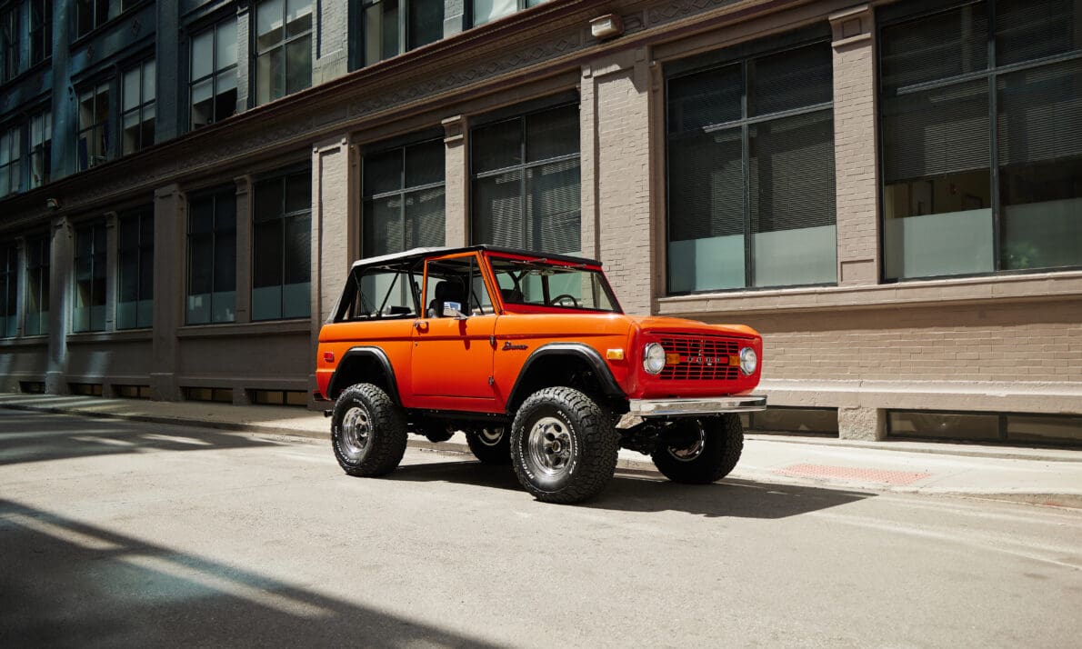 Classic Ford Bronco in bright orange parked on a city street, featuring lifted suspension, off-road tires, chrome wheels, and vintage styling with removable hardtop.